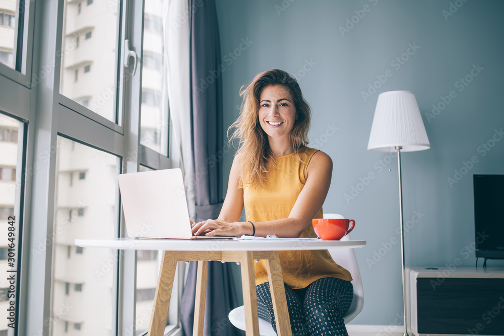 Portrait of happy housewife sitting at table in cozy apartment and spending leisure time for online shopping in web stores, positive female freelancer using home internet connection on laptop - obrazy, fototapety, plakaty 
