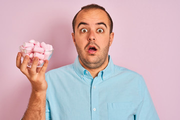 Sticker - Young man holding bowl with marshmallows standing over isolated pink background scared in shock with a surprise face, afraid and excited with fear expression