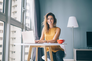 Portrait of happy housewife sitting at table in cozy apartment and spending leisure time for online shopping in web stores, positive female freelancer using home internet connection on laptop