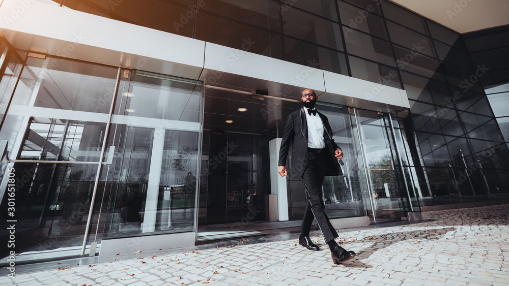 An elegant bearded bald African man entrepreneur is leaving a business skyscraper, he has just passed automatic sliding doors; a black businessman in a formal suit is leaving a modern building - obrazy, fototapety, plakaty 