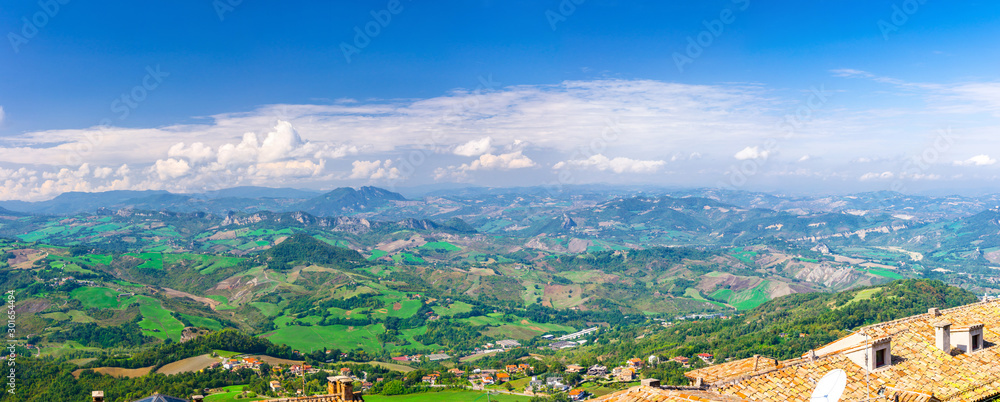 Obraz na płótnie Aerial top panoramic view of landscape with valley, green hills, fields and villages of Republic San Marino suburban district with blue sky white clouds background. View from San Marino fortress. w salonie