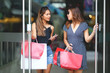 © NAMPIX - Two beautiful girls with sun glasses are holding shopping bags and smartphone walking out of the mall with happiness.