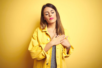 Poster - Young beautiful woman wearing denim jacket standing over yellow isolated background smiling with hands on chest with closed eyes and grateful gesture on face. Health concept.