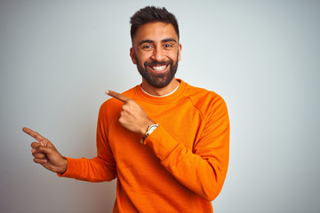 Wall Mural - Young indian man wearing orange sweater over isolated white background smiling and looking at the camera pointing with two hands and fingers to the side.