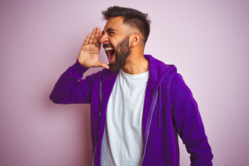 Wall Mural - Young indian man wearing purple sweatshirt standing over isolated pink background shouting and screaming loud to side with hand on mouth. Communication concept.