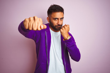 Wall Mural - Young indian man wearing purple sweatshirt standing over isolated pink background Punching fist to fight, aggressive and angry attack, threat and violence