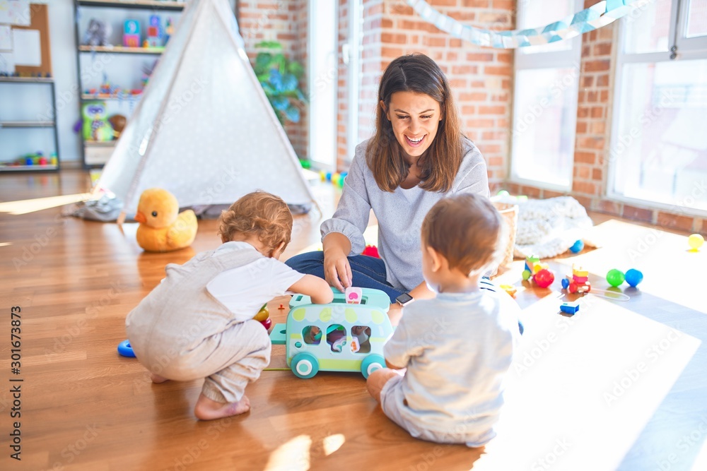 Beautiful teacher and toddlers playing around lots of toys at kindergarten - obrazy, fototapety, plakaty 