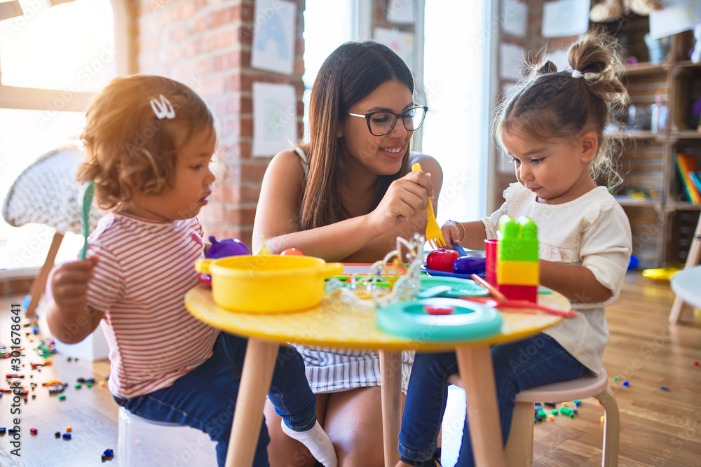 Young beautiful teacher and toddlers playing meals using plastic food and cutlery toy at kindergarten - obrazy, fototapety, plakaty 