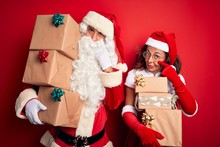 Middle Age Couple Wearing Santa Costume Holding Tower Of Gifts Over Isolated Red Background Pointing To The Eye Watching You Gesture, Suspicious Expression