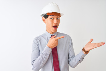 Wall Mural - Chinese architect man wearing tie and helmet standing over isolated white background amazed and smiling to the camera while presenting with hand and pointing with finger.