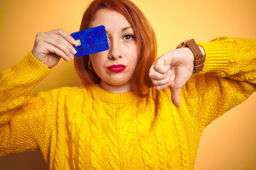 Wall Mural - Young beautiful redhead woman holding credit card over yellow isolated background with angry face, negative sign showing dislike with thumbs down, rejection concept