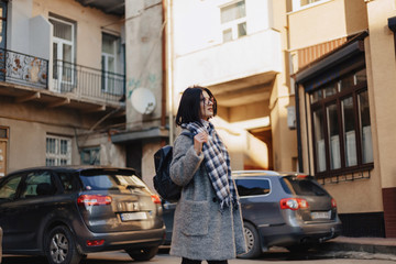 Wall Mural - Attractive positive young girl wearing glasses in a coat on the background of buildings on cars