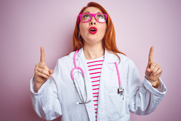 Canvas Print - Young redhead doctor woman using stethoscope standing over isolated pink background amazed and surprised looking up and pointing with fingers and raised arms.