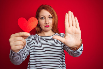 Poster - Beautiful redhead romantic woman holding heart standing over isolated red background with open hand doing stop sign with serious and confident expression, defense gesture