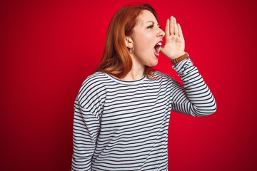 Sticker - Young redhead woman wearing strapes navy shirt standing over red isolated background shouting and screaming loud to side with hand on mouth. Communication concept.