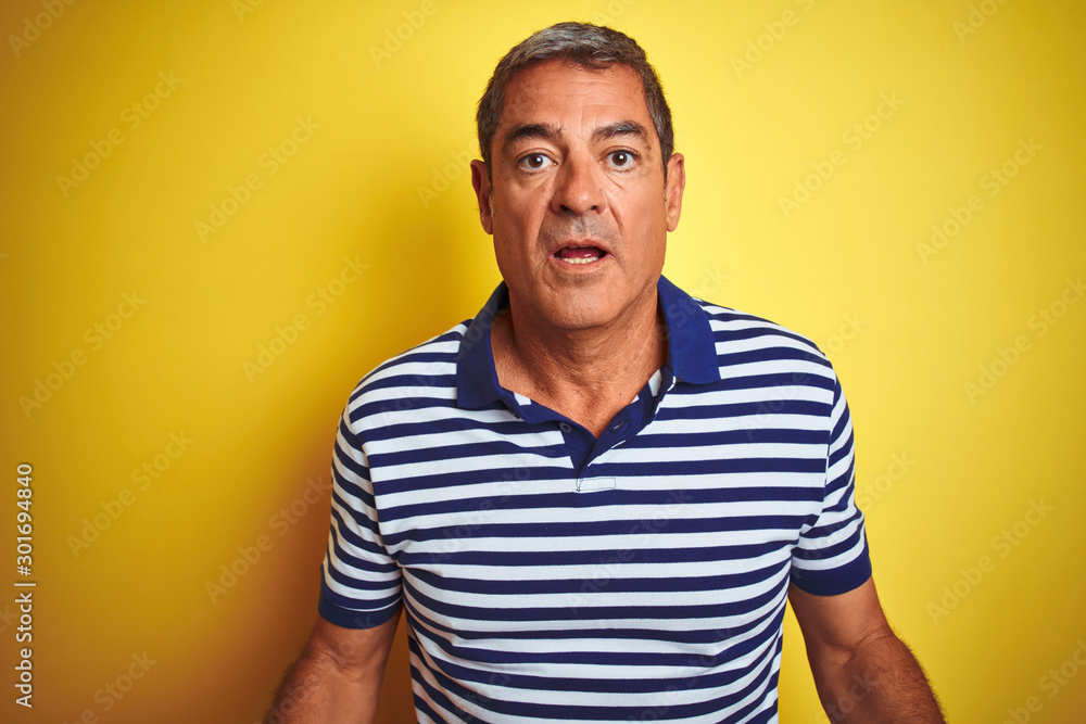 Handsome middle age man wearing striped polo standing over isolated yellow background afraid and shocked with surprise expression, fear and excited face. - obrazy, fototapety, plakaty 