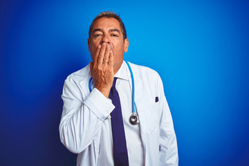 Wall Mural - Handsome middle age doctor man wearing stethoscope over isolated blue background bored yawning tired covering mouth with hand. Restless and sleepiness.