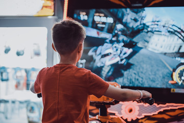 Wall Mural - Cute European boy in red t-shirt playing car racing on slot-machine. He enjoying his spare time. Back view.