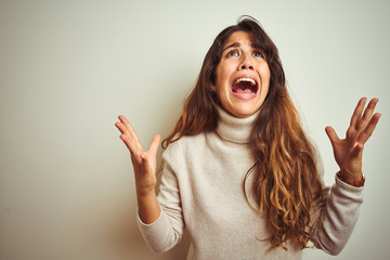 Wall Mural - Young beautiful woman wearing winter sweater standing over white isolated background crazy and mad shouting and yelling with aggressive expression and arms raised. Frustration concept.