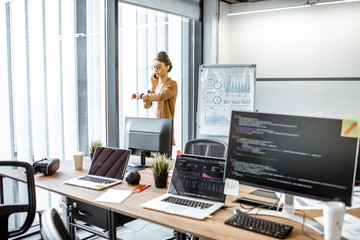 Wall Mural - Young business woman talking on the phone while standing near the window in the modern office with computers on the foreground