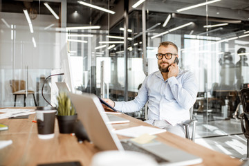 Senior confident manager talking on the phone while sitting at the working place in the modern office