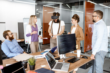 Wall Mural - Group of diverse colleagues during small conference in the office, creative bearded man trying a new product with virtual reality goggles