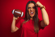Young beautiful sportswoman holding football ball over isolated red background annoyed and frustrated shouting with anger, crazy and yelling with raised hand, anger concept