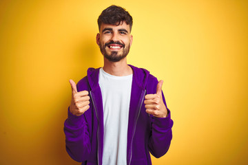 Young man with tattoo wearing sport purple sweatshirt over isolated yellow background success sign doing positive gesture with hand, thumbs up smiling and happy. Cheerful expression