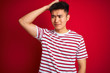 © Krakenimages.com - Young asian chinese man wearing striped t-shirt standing over isolated red background confuse and wonder about question. Uncertain with doubt, thinking with hand on head. Pensive concept.