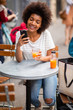 © mimagephotos - beautiful young black woman sitting at outdoor cafe with mobile phone and drink