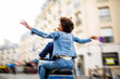 © mimagephotos - behind of young african american woman sitting on back of scooter driving through city streets with arms raised