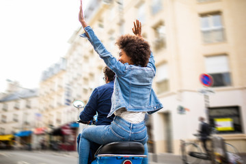 behind of happy young african american woman sitting on back of scooter driving through city streets with arms raised