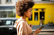© mimagephotos - Side of beautiful young black woman with afro hairstyle walking with cellphone in city
