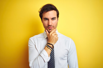 Poster - Young handsome businessman wearing elegant shirt and tie over isolated yellow background looking confident at the camera with smile with crossed arms and hand raised on chin. Thinking positive.