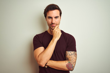 Wall Mural - Young handsome man with tattoo wearing purple casual t-shirt over isolated white background looking confident at the camera with smile with crossed arms and hand raised on chin. Thinking positive.