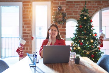 Poster - Beautiful woman sitting at the table working with laptop at home around christmas tree touching mouth with hand with painful expression because of toothache or dental illness on teeth