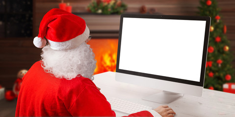 Santa Claus work on computer beside fireplace and Christmas tree. Christmas greeting card concept with isolated computer display for greeting text.
