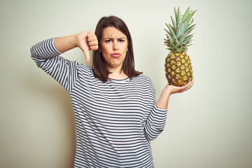 Sticker - Young beautiful woman holding tropical fruit pineapple over isolated background with angry face, negative sign showing dislike with thumbs down, rejection concept