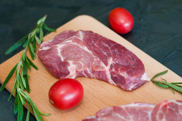 Wall Mural - Raw pieces of pork, rosemary and tomatoes on a cutting board, on dark gray concrete background, close-up