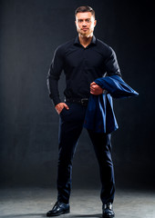 Wall Mural - Fashion portrait of young man in black shirt poses over dark wall with one hand in pocket. Jacket in a hand. Handsome confident man, businessman.
