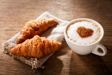 Wall Mural - Valentines day.  Cup of cappuccino coffee with drawn heart and fresh croissants, top view