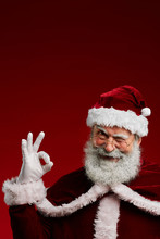 Waist Up Portrait Of Winking Santa Claus Looking At Camera And Showing OK Sign While Posing Against Red Background, Copy Space