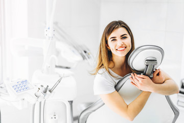 Wall Mural - Portrait of a happy young patient after the dental examination. A woman sitting hugging armchair in withe modern dentist office