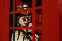 Outdoor Close Up Portrait Of Young Elegant Woman Wearing Trendy White Cat Eye Sunglasses, Leather Beret, Posing In Red Phone Booth, Talking On The Phone. Copy, Epmty Space For Text