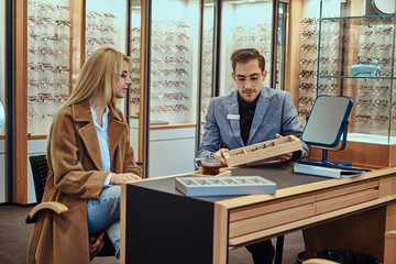 Wall Mural - Woman is choosing eyeglasses while male optician sitting near with another eyeglasses.