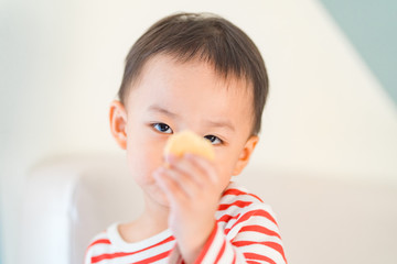 Wall Mural - Cute little asian toddler boy holding cookie and eating cookie in sofa at home.Hungry toddler boy make delicious face when he eating cookies.