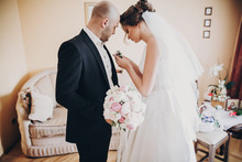 Beautiful Bride Putting Stylish Boutonniere On Groom Suit, Standing In Room. Gorgeous Woman In White Gown And Man In Suit Getting Ready For Wedding Ceremony