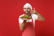 Male chef cook or baker man in striped apron toque chefs hat posing isolated on red background. Cooking food concept. Mock up copy space. Hold plate with salad, raised hand to face feeling food smell.