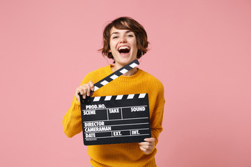 Wall Mural - Laughing young brunette woman girl in yellow sweater posing isolated on pastel pink background in studio. People lifestyle concept. Mock up copy space. Holding classic black film making clapperboard.