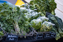 Close-up Of Bundles Of Leafy Green Vegetables Stacked For Sale At A Farmers Market. Varieties Of Kale, And Arugula And Asian Spinach.